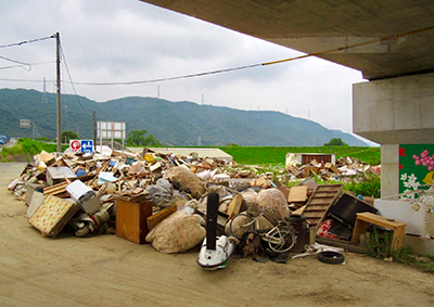 西日本豪雨に襲われた岡山県真備町で、川べりに積まれている災害ごみ。（写真：ラジオ関西）