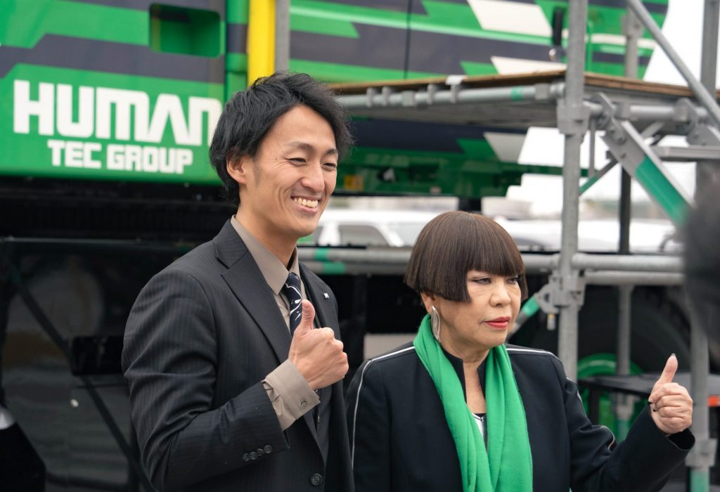 クレーン車の前でポーズを取るコシノジュンコさん（右）と濱中朋弘さん（左）